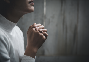 Person in a white sweater sitting with clasped hands, appearing contemplative or in prayer, against a wooden background.