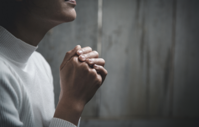 Person in a white sweater sitting with clasped hands, appearing contemplative or in prayer, against a wooden background.