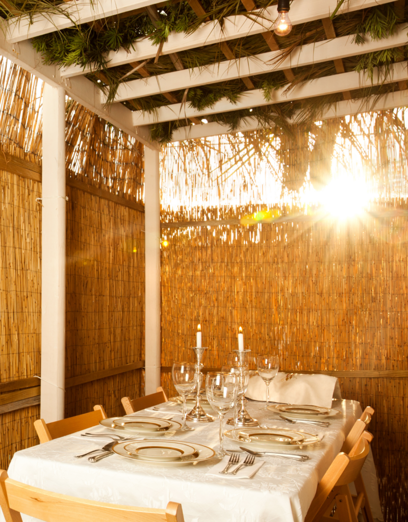 Outdoor dining space with a set table for six, two lit candles, and sunlight streaming through bamboo walls and roof.