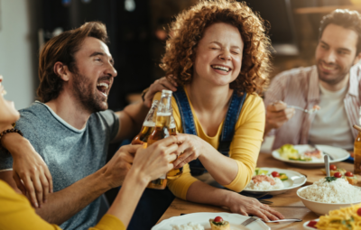 Group of friends laughing and clinking beer bottles at a dining table with plates of food.