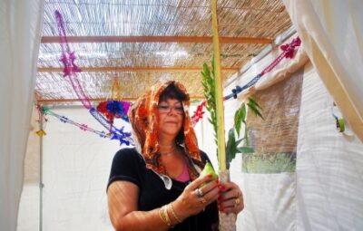 A woman holding a lulav and etrog in a Sukkah decorated with colorful streamers.