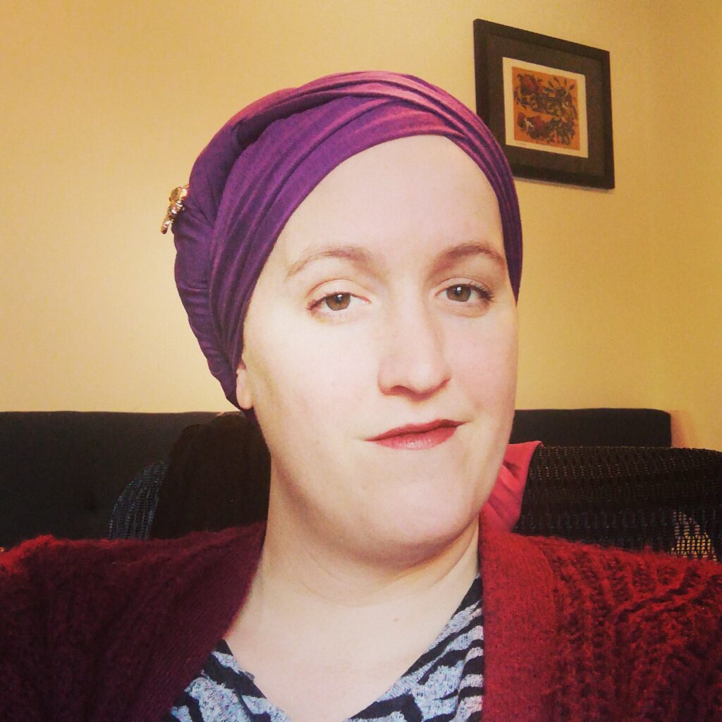 Person wearing a purple headscarf and maroon sweater, sitting indoors with a framed picture on the wall behind.