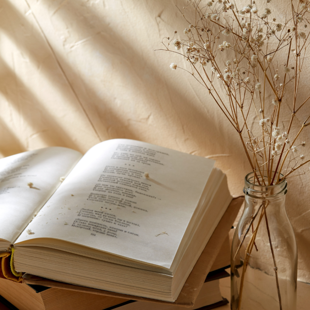 Open book with poetry on a table, next to a clear vase holding dried flowers. Soft sunlight casts shadows.