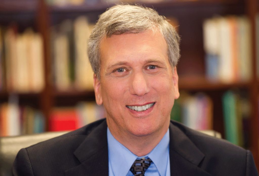 Smiling man in a suit and tie in front of a blurred bookshelf background.