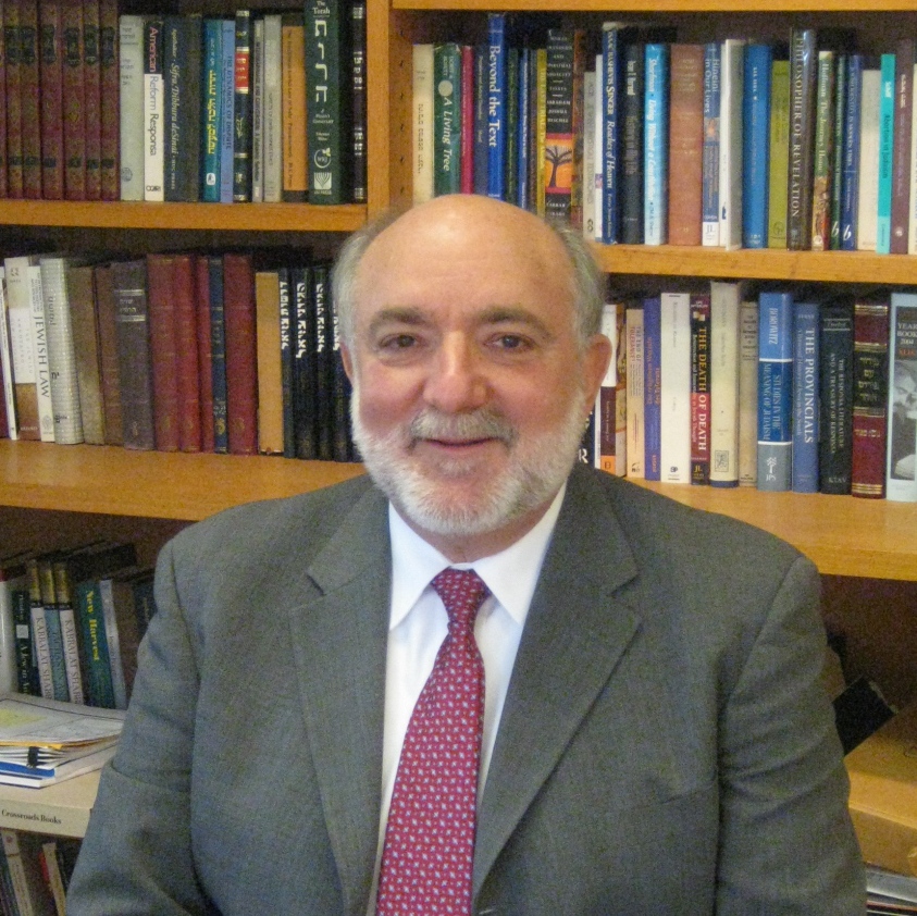 A bearded man in a suit sits in front of bookshelves filled with various books.