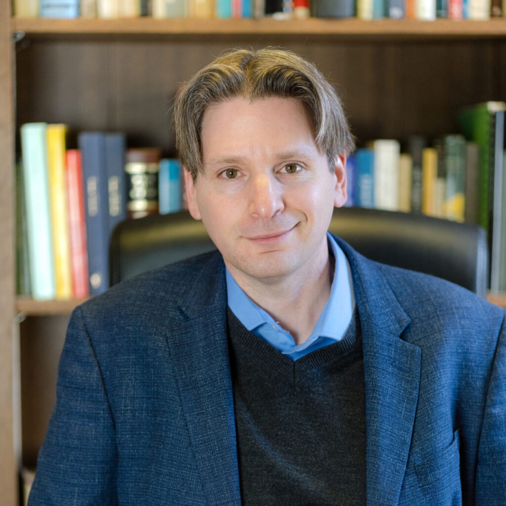 A person in a blue blazer sits in front of a bookshelf, smiling.