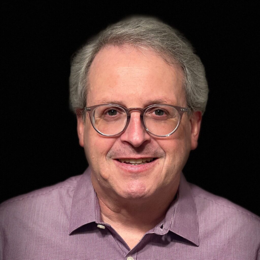A man with glasses and a purple shirt smiling against a black background.