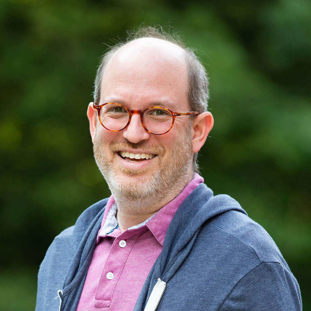 Smiling man with glasses, wearing a pink shirt and blue hoodie, standing outdoors against a blurred green background.