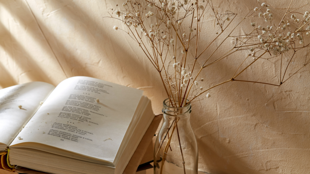 Open book on a table next to a glass vase holding dried flowers against a beige textured wall.