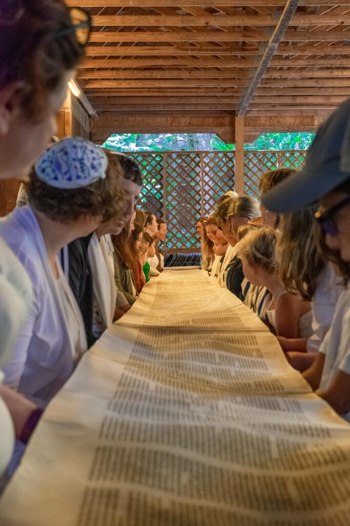 A group of people gathered around a table, reading a long scroll in a wooden pavilion.
