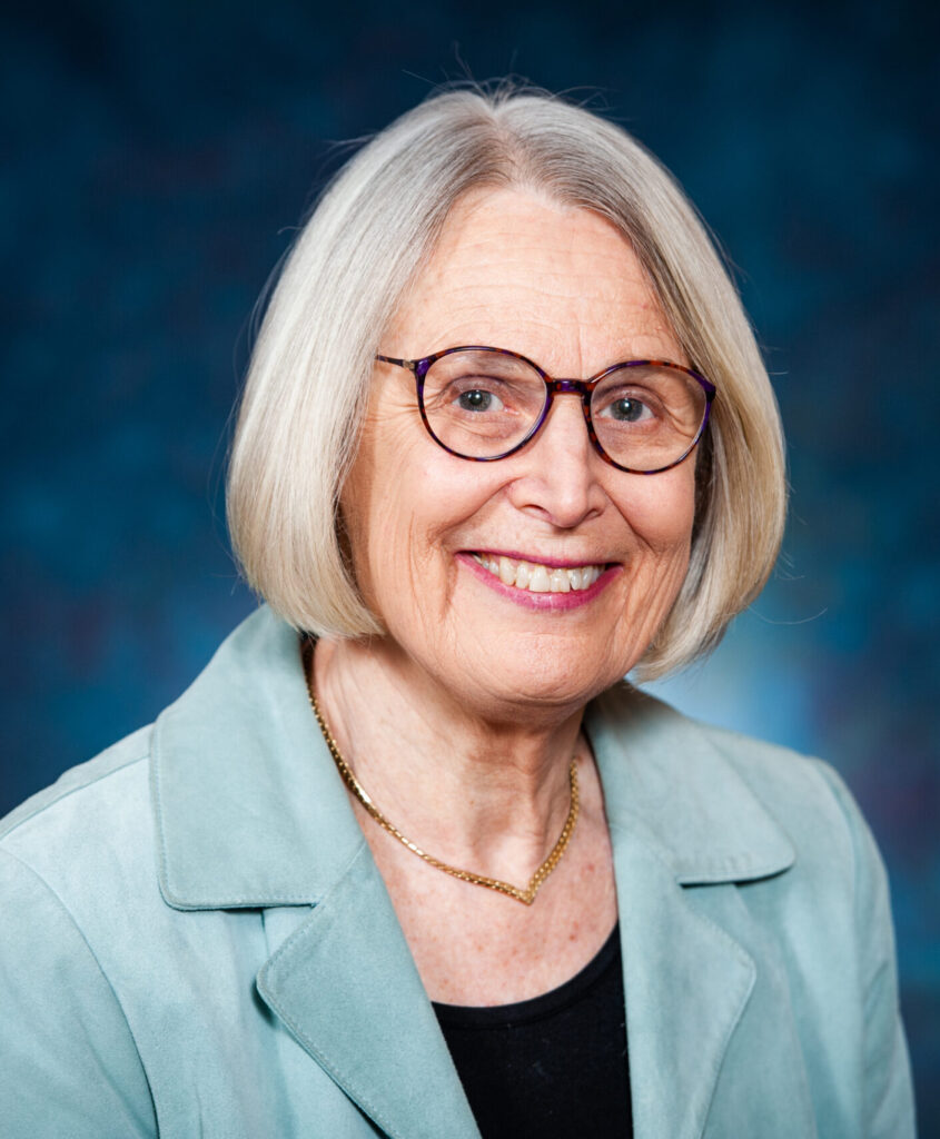 Smiling woman with gray hair, glasses, light blue blazer, and black top against a blue background.