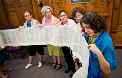 A group of people holding and reading a long scroll, standing together in a room with wooden walls.