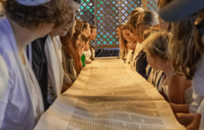 Multiple people gathered around either side of a long table, the Torah unrolled onto the table