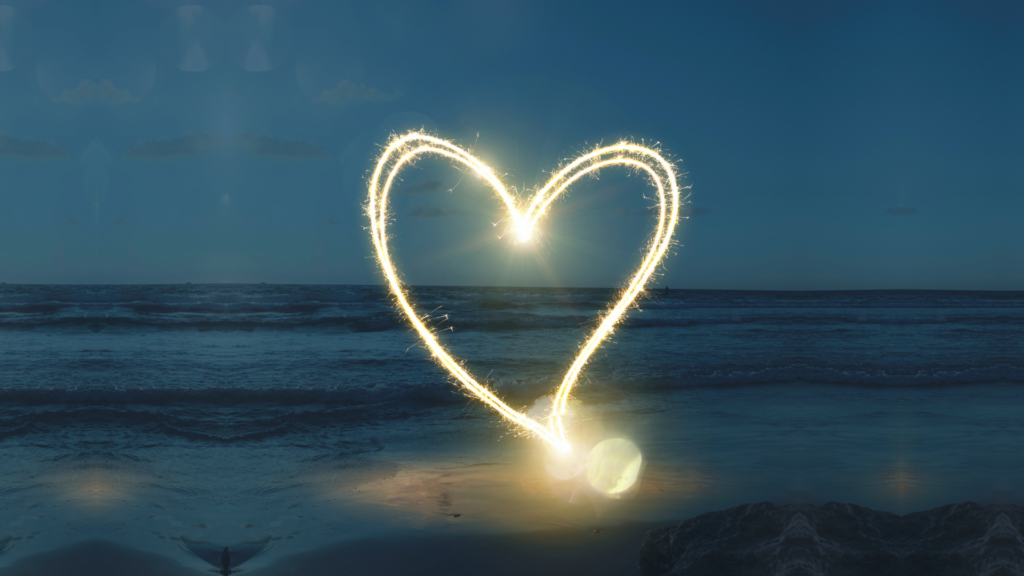 An illuminated, floating heart on a beach, with ocean water in the background. This image was to promote the recent Ritualwell program.