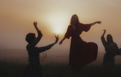 Silhouettes of three people dancing in a field at sunset, with the sky glowing softly in the background.