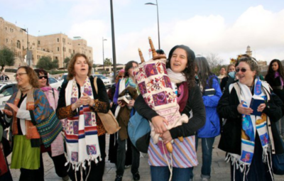 Group of women walking outdoors, one holding a Torah scroll, others clapping and smiling, with buildings in the background.