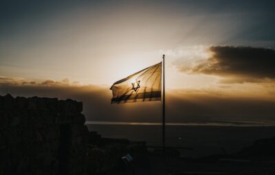 Israeli flag waves against a dramatic sunset, silhouetted by clouds and distant landscape.