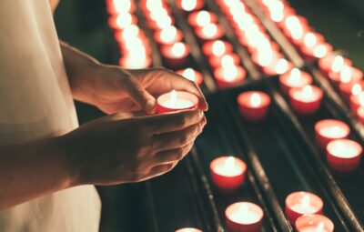 Person holding a lit candle among rows of burning candles in a dimly lit setting.