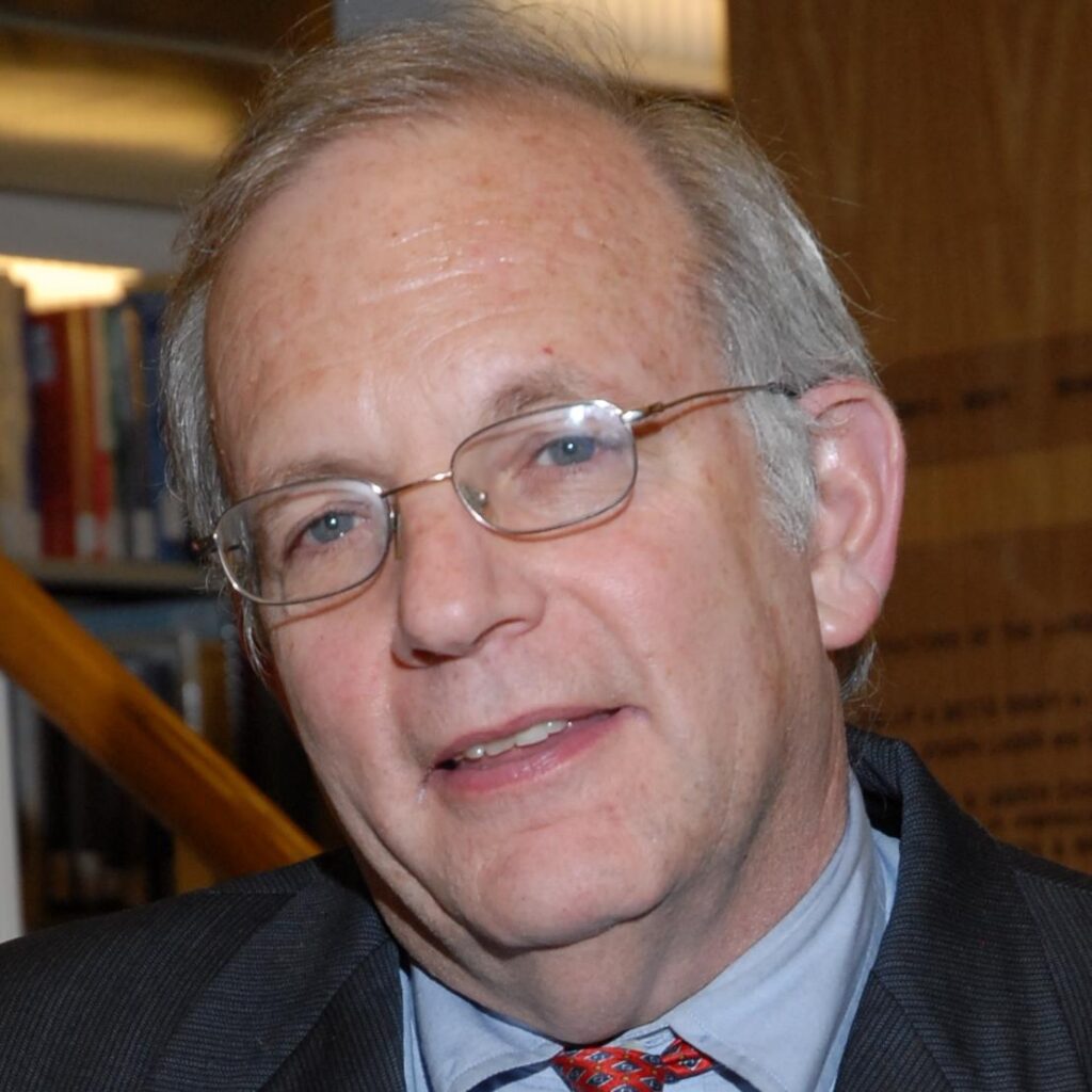 A person wearing glasses, a suit, and tie, standing indoors with shelves in the background.