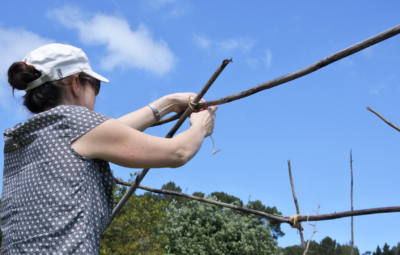 A woman in a cap ties sticks together outdoors under a blue sky.