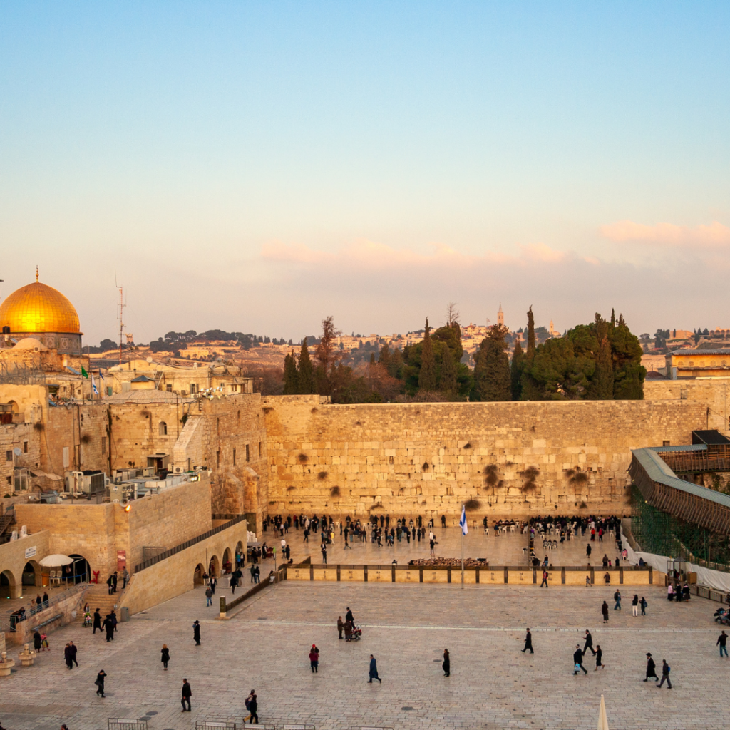 View of the Western Wall plaza with people gathered, and the Dome of the Rock visible under a clear sky.