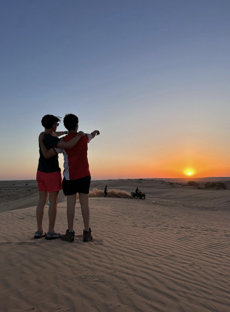 Two people embracing and pointing at a sunset over sand dunes, with an ATV kicking up dust in the background.