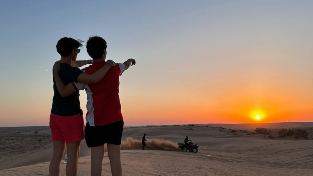 Two people stand arm in arm, watching a sunset in the desert while riders on ATVs drive across the sand dunes.