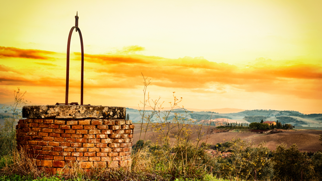 A rustic well with a metal handle in a scenic countryside at sunset, overlooking hills and distant farms.