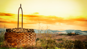 A rustic well with a metal handle in a scenic countryside at sunset, overlooking hills and distant farms.