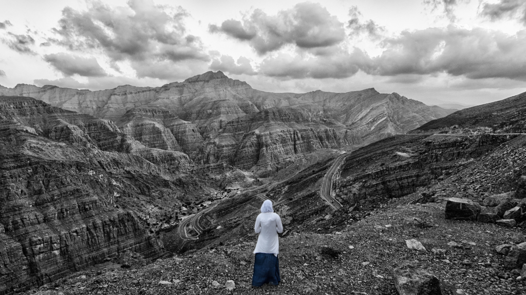 Person in white stands on a rocky hill overlooking a dramatic mountain landscape under a cloudy sky.