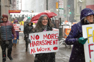 A rabbi holds a "no more harrasment at work" sign on the streets of Philadelphia. It's snowing and people around her are carrying umbrellas. Her hair is dotted with white snowflakes.