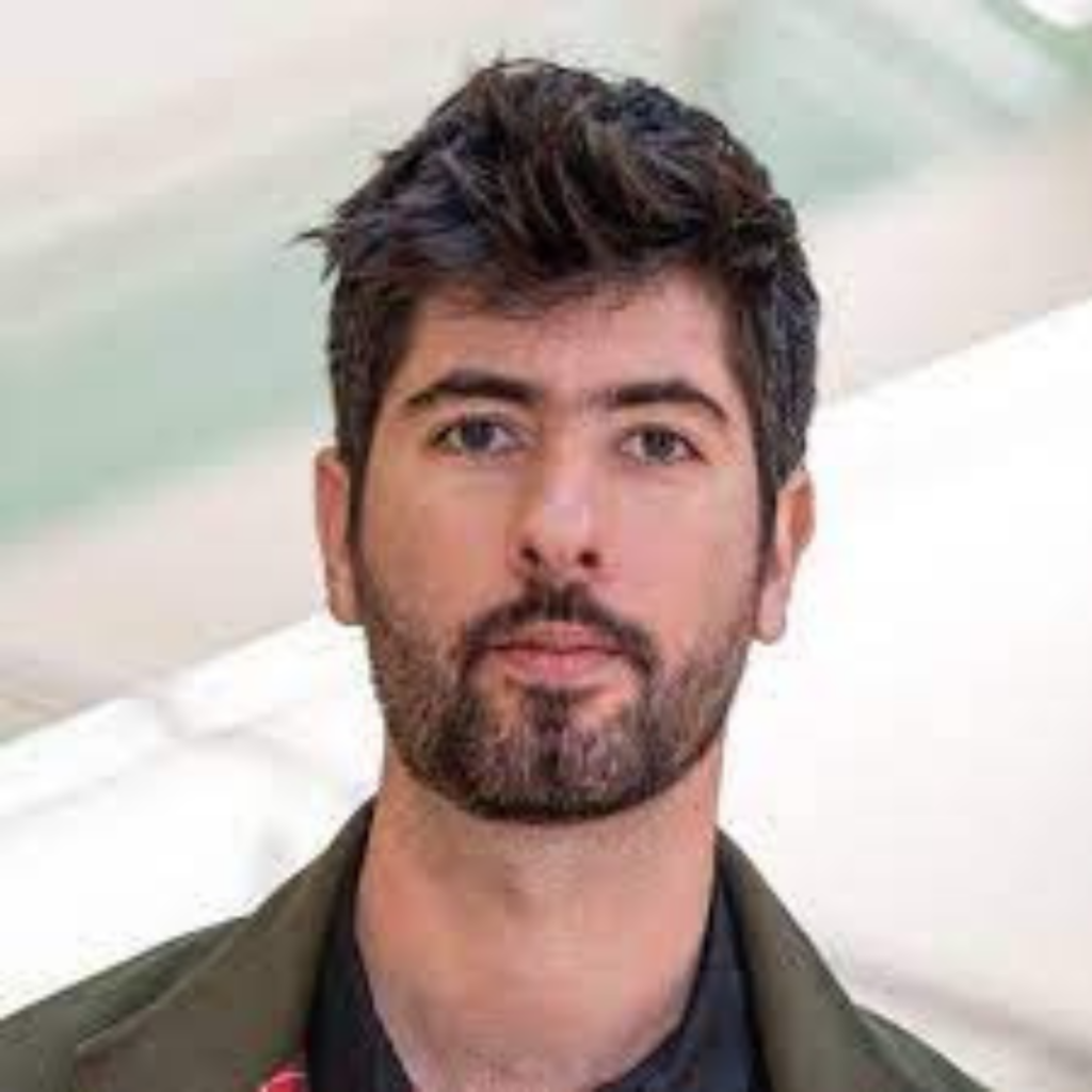 A person with dark hair and a beard is wearing a jacket, indoors.