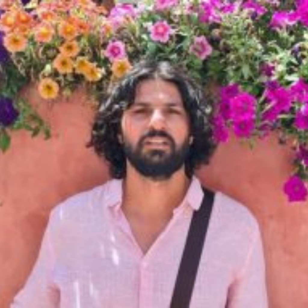 Man with curly hair in pink shirt in front of a wall with colorful hanging flowers.