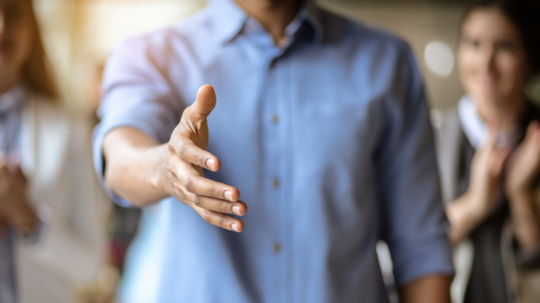 A person extending their hand for a handshake, with a blurred background and people clapping.