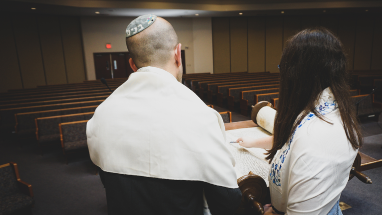 Two people in a synagogue facing an empty pew, one holding a Torah scroll.