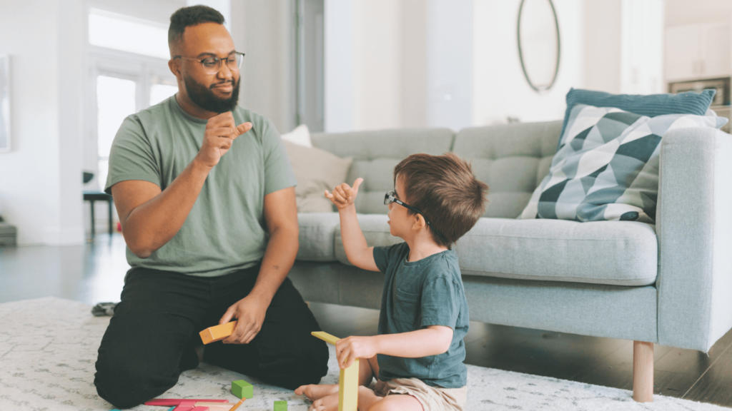 Black man signing in American Sign Language with a little white boy in a living room