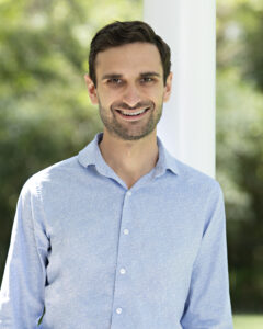 A smiling man in a light blue shirt stands outdoors with greenery in the background.