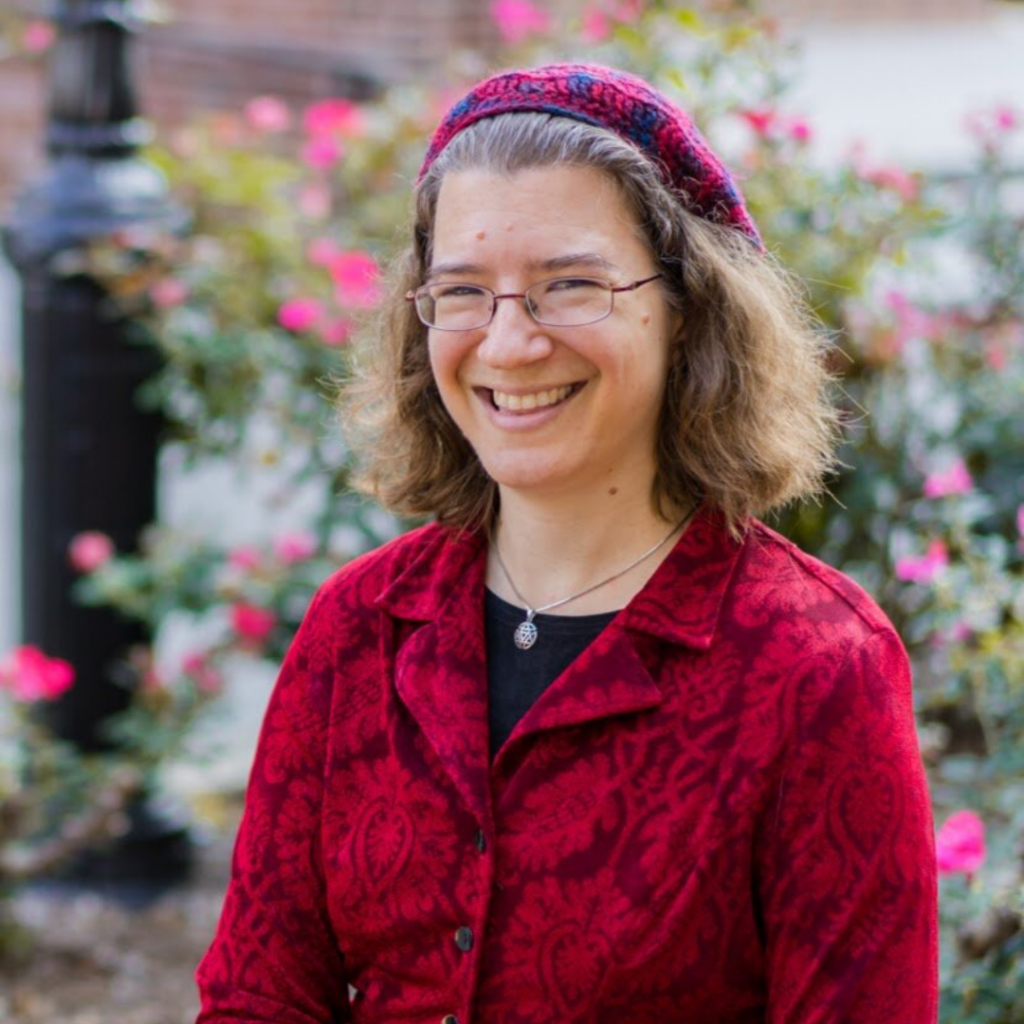 A person with glasses and a red patterned headscarf smiles in front of pink flowers.