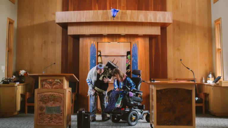 A person in a wheelchair holding a Torah scroll with another person helping in a synagogue.