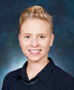 Person smiling against a blue backdrop, wearing a dark shirt and a short hairstyle.