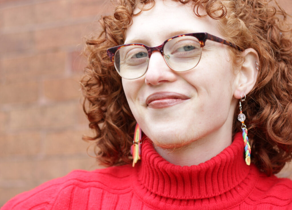 Person with curly hair, glasses, and colorful earrings smiles, wearing a red turtleneck, against a brick wall background.