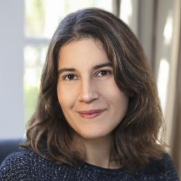 A person with shoulder-length brown hair and a slight smile, sitting indoors with light coming from a window.