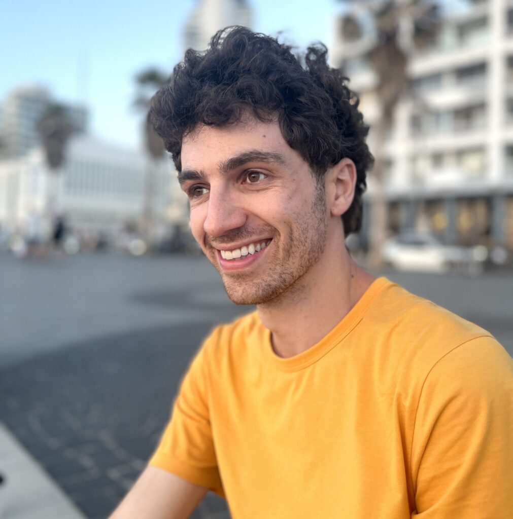 Young man in a yellow shirt smiling outdoors, with a blurry urban background.