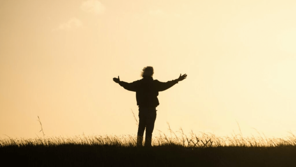 Person with their arms open in a field silhouetted against a yellow sky