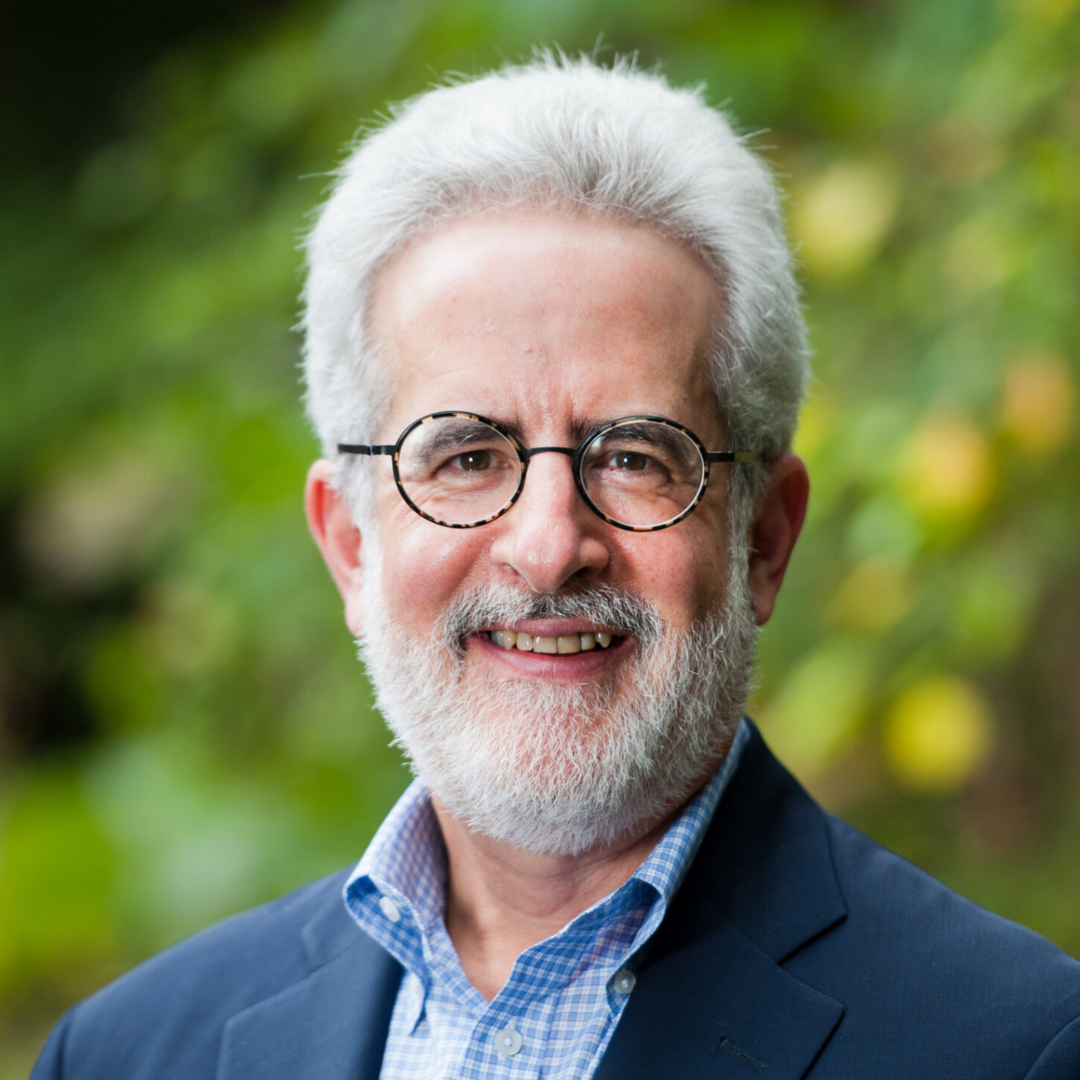 A smiling man with glasses and a white beard, wearing a suit jacket and blue shirt, set against a blurred green background.
