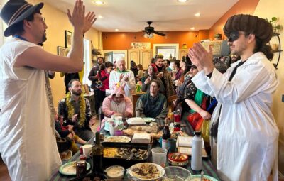 People in festive attire gather indoors, some in costumes, clapping near a table filled with food and drinks.