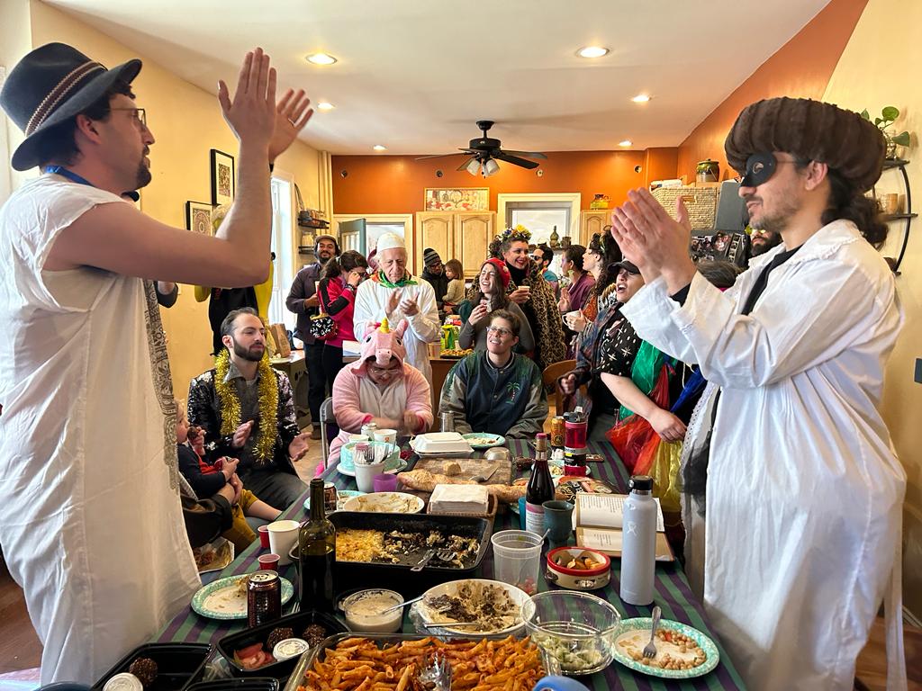 People in festive attire gather indoors, some in costumes, clapping near a table filled with food and drinks.