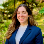 Person in a blue blazer smiling outdoors with greenery in the background.