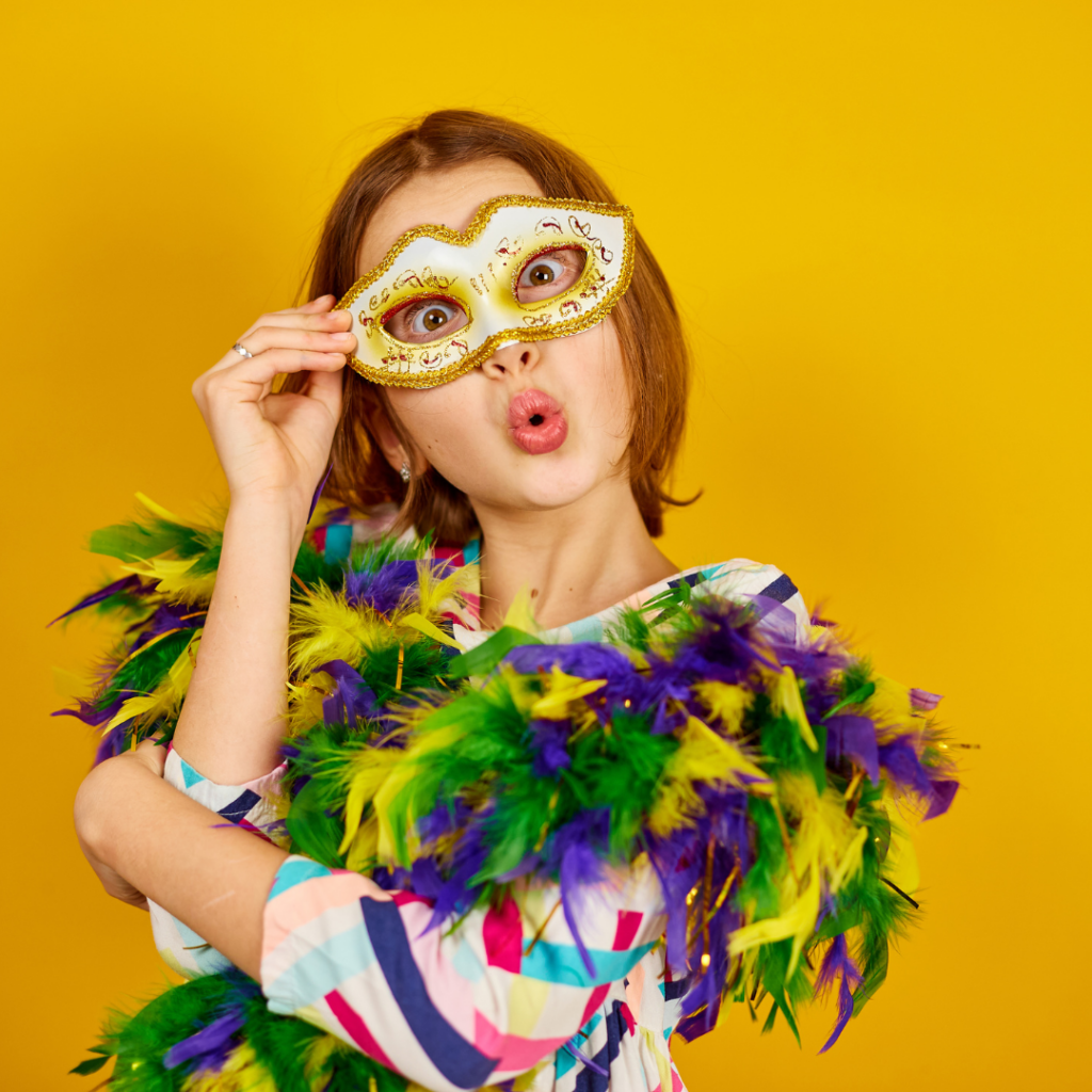 Person in a colorful outfit and feather boa poses playfully with a decorative masquerade mask against a yellow background.