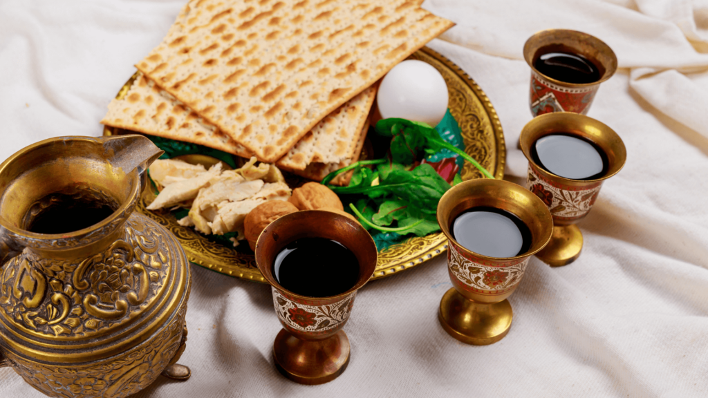 A seder plate and four cups of red wine in gold wine glasses, next to a pitcher of red wine
